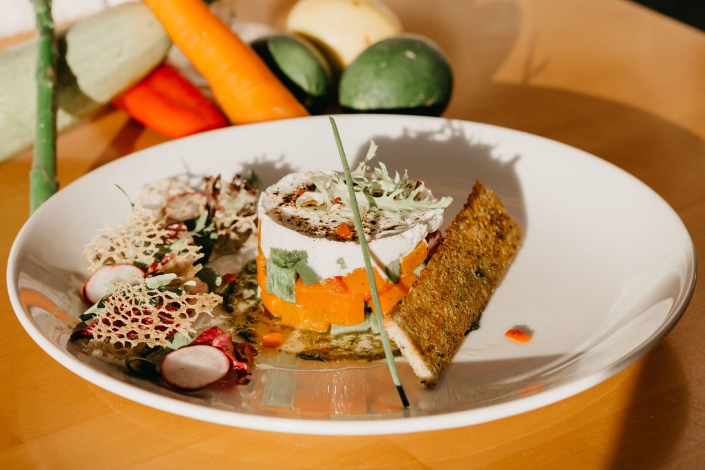 High angle of healthy vegetable tartar served on white ceramic plate with salad and crispy bread near scattered fresh ingredients in kitchen in sunlight